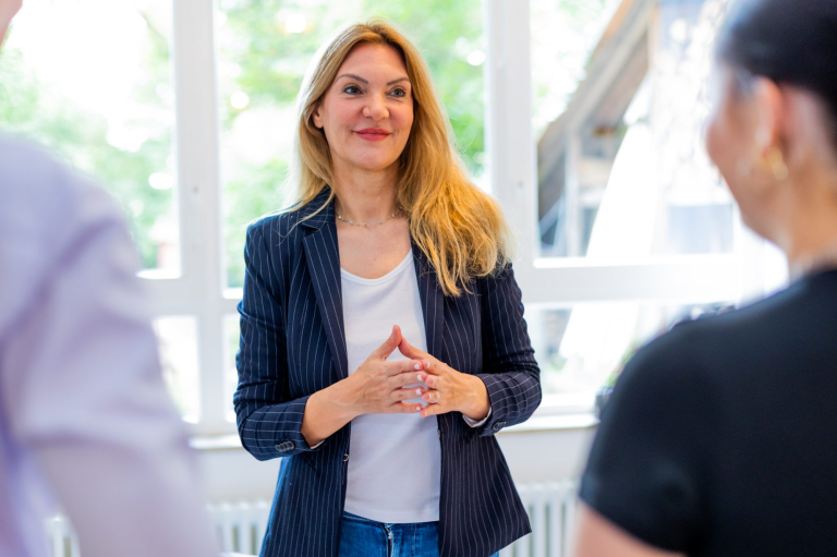 Frau mit brunetten Haaren, in Blazer, spricht mit zwei Personen in einem hellen Raum.